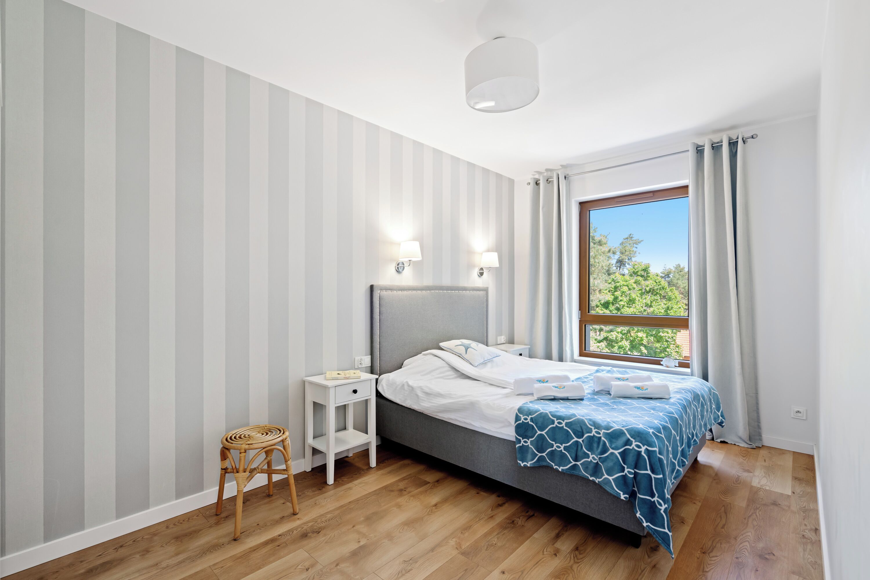A bedroom with a bed, bedside tables, a chair, a window with curtains, and a view of greenery.