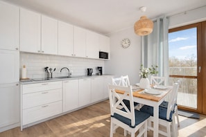 A modern kitchen with white cabinets, a wooden dining table, and a view of the outdoors through a large window.