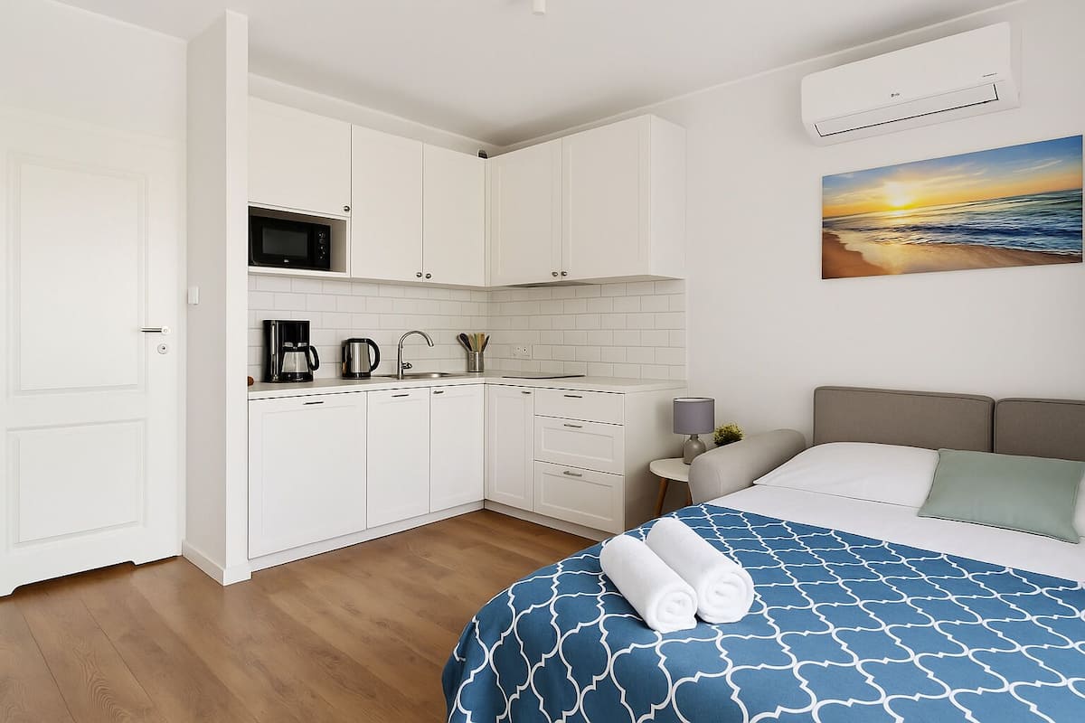 A compact room with a kitchenette, a bed with a patterned blue and white bedspread, and a painting of a sunset on the wall.
