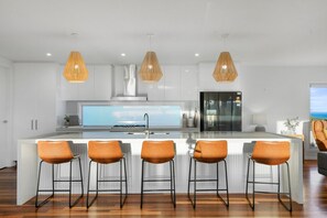 A modern kitchen with a central island, bar stools, pendant lights, and a large window overlooking the ocean.