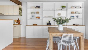 Une cuisine moderne avec des meubles blancs, une table à manger en bois et des chaises blanches. On y trouve une étagère blanche ornée d’objets décoratifs et surmontée d’une plante en pot.