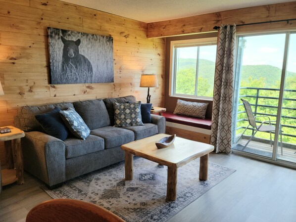 A cozy room with a wooden wall, a sofa with patterned cushions, a small table, a window with a view of greenery, and a wooden floor. - Mountain Villa Magnificent Views - 2 Pools, Hot Tub & Fireplace (Gatlinburg)