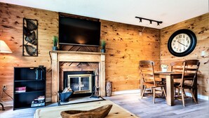 A cozy living room with a fireplace, wooden walls, a dining table with chairs, a large wall clock, and a flat-screen TV mounted above the fireplace.