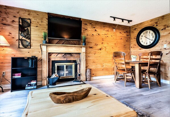 A cozy living room with a fireplace, wooden walls, a dining table with chairs, a large wall clock, and a flat-screen TV mounted above the fireplace.