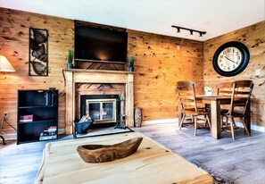 Un salon chaleureux avec une cheminée, des murs en bois, une table à manger avec des chaises, une grande horloge murale et un téléviseur à écran plat fixé au-dessus de la cheminée.