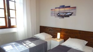 A hotel room with two beds, a wooden headboard, a nightstand with a lamp, and a window with curtains.