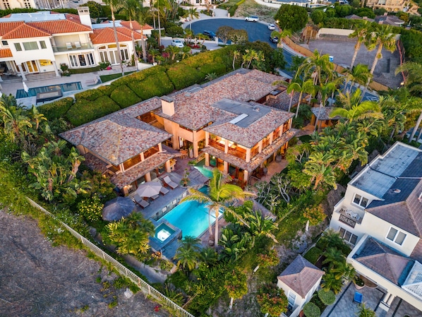 A large, multi-story house with a swimming pool, surrounded by lush greenery and palm trees.