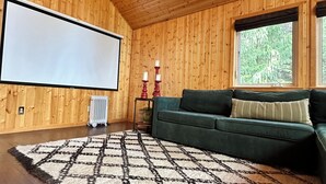 A wooden-paneled room with a projector screen, a green sofa, a patterned rug, and a radiator. - Sierra City Loves Falls Cabin - Walking Distance to Waterfall & PCT (Sierra City)