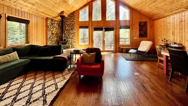 A log cabin interior with a stone fireplace, wooden walls, and large windows. - Sierra City Loves Falls Cabin - Walking Distance to Waterfall & PCT (Sierra City)