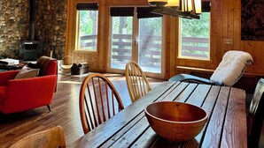 A cozy cabin interior with a stone fireplace, wooden ceiling, large windows, and a dining table. - Sierra City Loves Falls Cabin - Walking Distance to Waterfall & PCT (Sierra City)