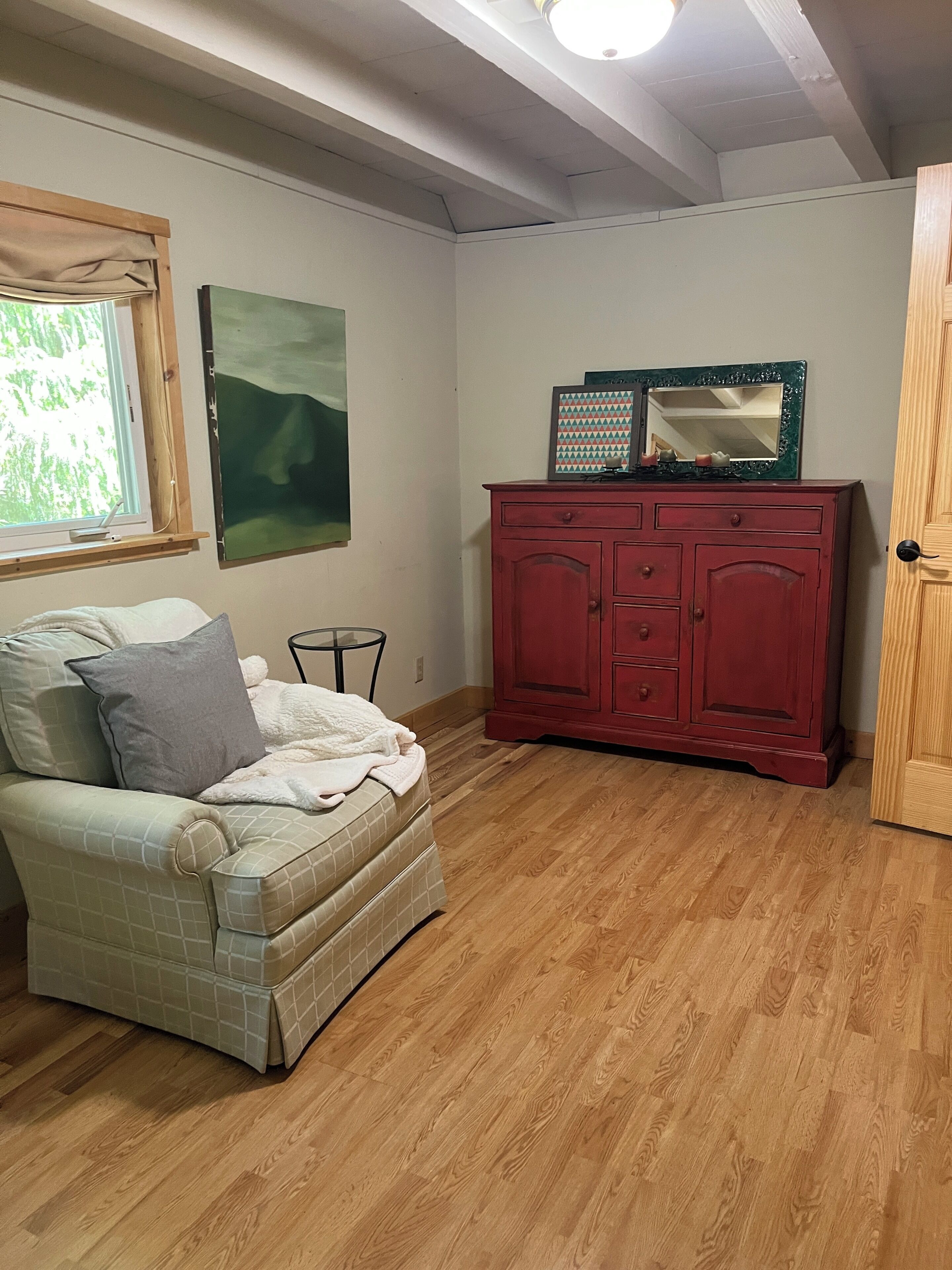 Une pièce avec un plancher en bois, un meuble rouge, une chaise blanche recouverte d’une couverture, une petite table et une fenêtre donnant sur un paysage verdoyant.