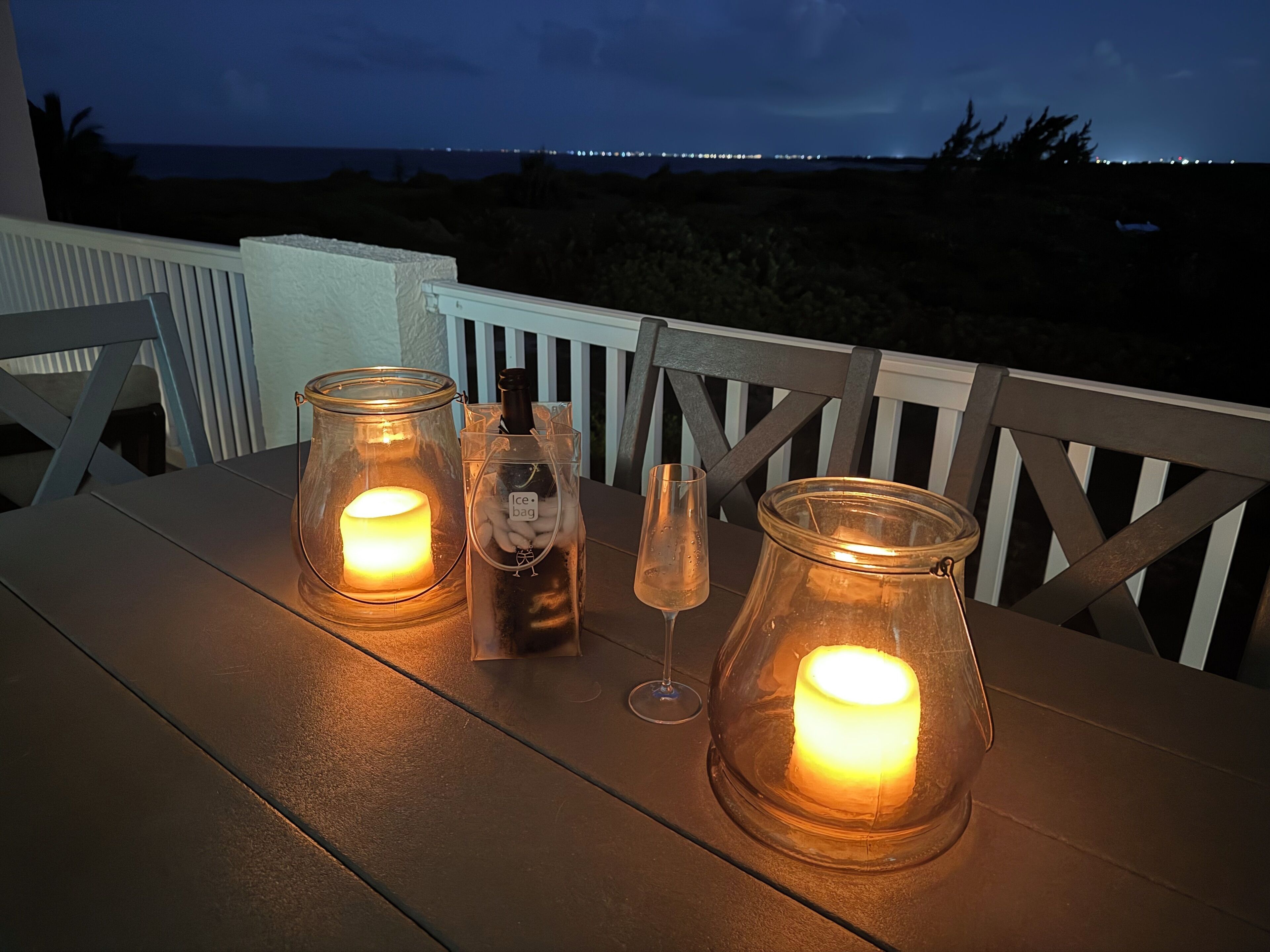 A table with two glass candle holders, a bottle, and a glass on a wooden deck overlooking a beach at night.