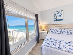 A hotel room with a bed, a nightstand, a lamp, a painting, and a view of the beach.