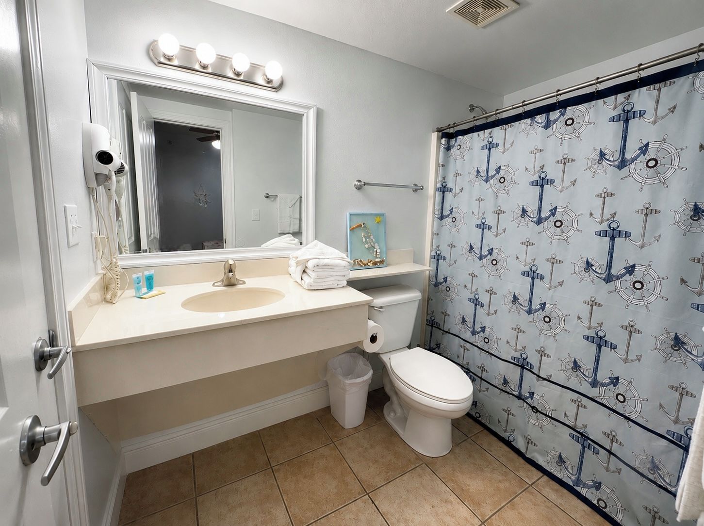 Un baño con tocador, inodoro, cortina de ducha con diseño de anclas y un espejo con luces superiores.