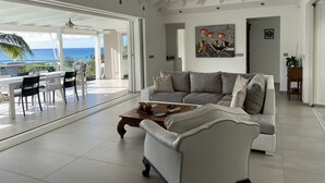 A spacious living room with a large gray sofa, a wooden coffee table, and a ceiling fan. There is a dining area with a table and chairs, and a view of the ocean through large glass doors. -  MAGNIFIQUE VILLA KYLA'S  the most beautiful sea view in the Caribbean with POOL (Collectivity of Saint Martin)
