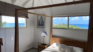 A bedroom with a wooden canopy bed, a nightstand with a lamp, a framed picture on the wall, and a view of the ocean through large windows. -  MAGNIFIQUE VILLA KYLA'S  the most beautiful sea view in the Caribbean with POOL (Collectivity of Saint Martin)