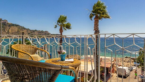 Eine Terrasse mit Korbmöbeln, einem Tisch mit einem Glas Rotwein und Blick auf das Meer und die felsige Küste.