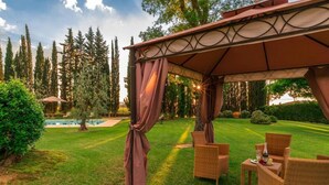 Un área de estar techada al aire libre con mobiliario de mimbre, piscina y un césped cuidado.