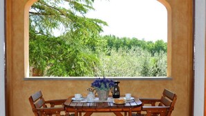 A dining table with chairs, a pitcher, cups, and a basket with flowers, set against a window with a view of trees and greenery. - Santa Margherita - NINFEA (Poggibonsi)