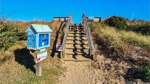 Un escalier en bois mène à l’accès à la plage. On y trouve une boîte aux lettres et un panneau sur le côté gauche.