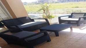 Un patio con mobiliario de mimbre, incluyendo sofás y una mesa, y vista a un paisaje verde a través del ventanal.