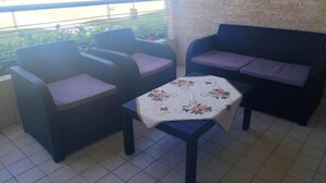 Un balcón con mobiliario de mimbre, una mesa con mantel floreado y vista a follaje y una masa de agua.