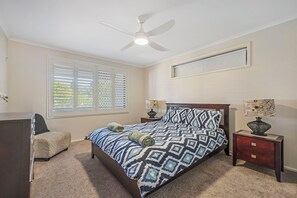 Un dormitorio con cama, mesita de noche, cómoda, ventilador de techo y ventana con persianas.