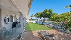 Eine Terrasse mit Glastisch und Stühlen, einem Grill und Blick auf ein Gewässer, umgeben von einem Zaun und Bäumen.