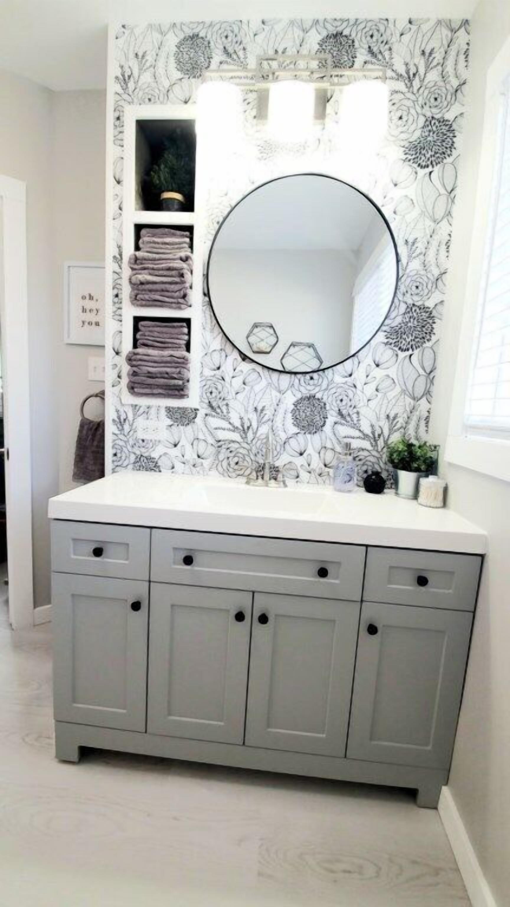 Un baño con tocador gris, un espejo redondo y papel tapiz con motivos florales.