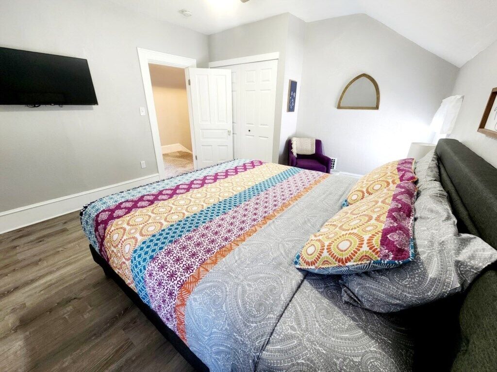 Un dormitorio con una cama, un televisor montado en la pared, una puerta y un pequeño rincón de descanso con una silla y una mesa.