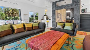 Un salon moderne avec un canapé et un pouf, une grande fenêtre donnant sur des palmiers, et un coin cuisine agrémenté d’un miroir et d’une suspension.