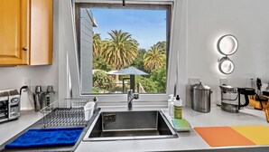 Une cuisine avec une fenêtre donnant sur des palmiers, un évier, un égouttoir, une machine à café et un grille-pain.