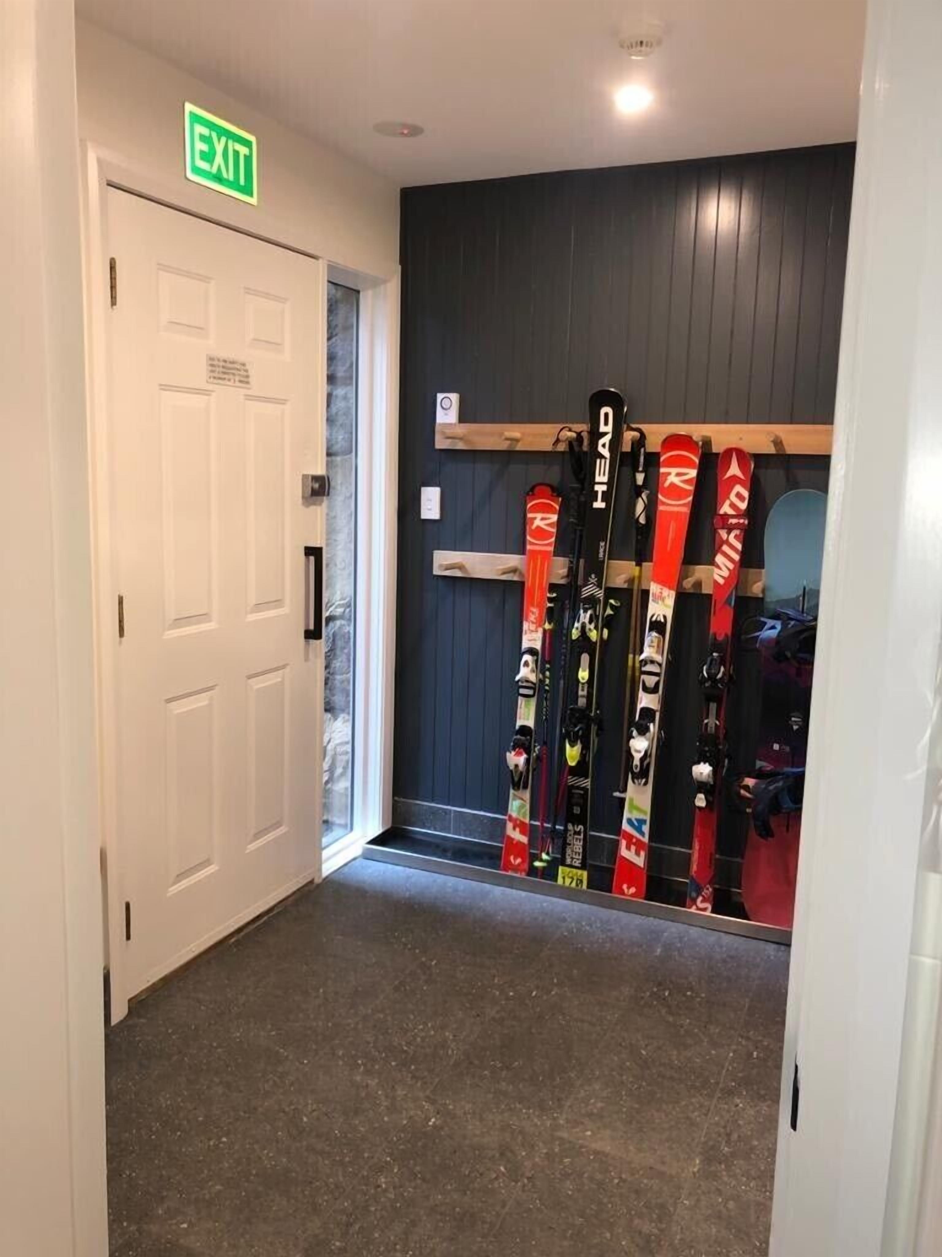 A hallway with an exit sign, a door, and a row of skis leaning against a wall.