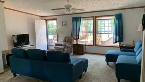 Ein Wohnzimmer mit blauem Sofa, einem Fernseher, einem Deckenventilator und großen Fenstern mit Vorhängen.