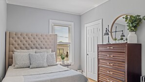 Un dormitorio con cabecera capitonada, una cama grande con ropa de cama estampada, un aparador con varios cajones y una ventana con persianas.