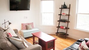 Un salón con un sofá gris, una mesa de centro roja, un sillón azul claro, un televisor montado en la pared y una estantería de madera con libros y objetos decorativos.