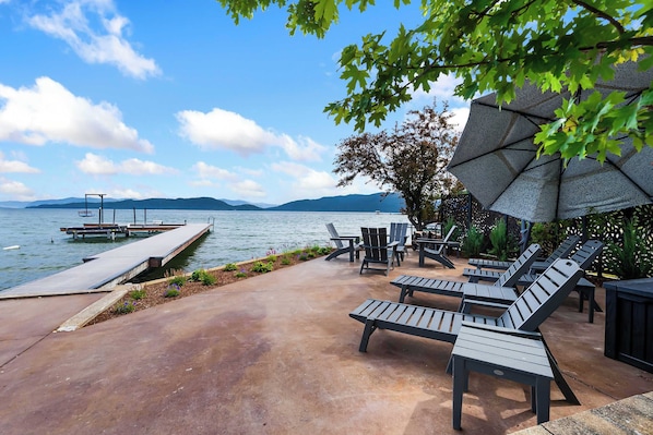 A waterfront patio with lounge chairs, a dock, and a large umbrella.