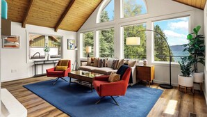 A spacious living room with a high ceiling, large windows, a sofa, a coffee table, and a red armchair. - Dog-Friendly Beachfront Getaway, Hot Tub, Boat Lift (Sandpoint)