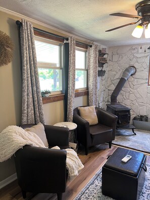 A cozy living room with two leather armchairs, a small round table, a stone wall, a wood-burning stove, and a ceiling fan.