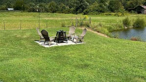 A grassy outdoor area with a fire pit and chairs, a small pond, and a mountain in the background.