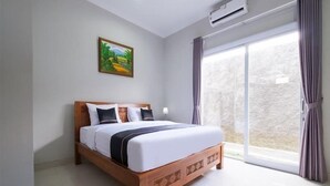 A hotel room with a bed, a painting on the wall, an air conditioning unit, and a view through a window with curtains. - OYO 3906 Dalem Putu Sanjaya Syariah (Sleman)