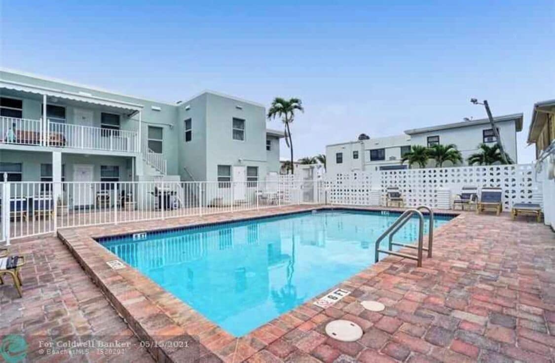 Una piscina con borde de ladrillo, rodeada por una valla blanca y un edificio con balcones.