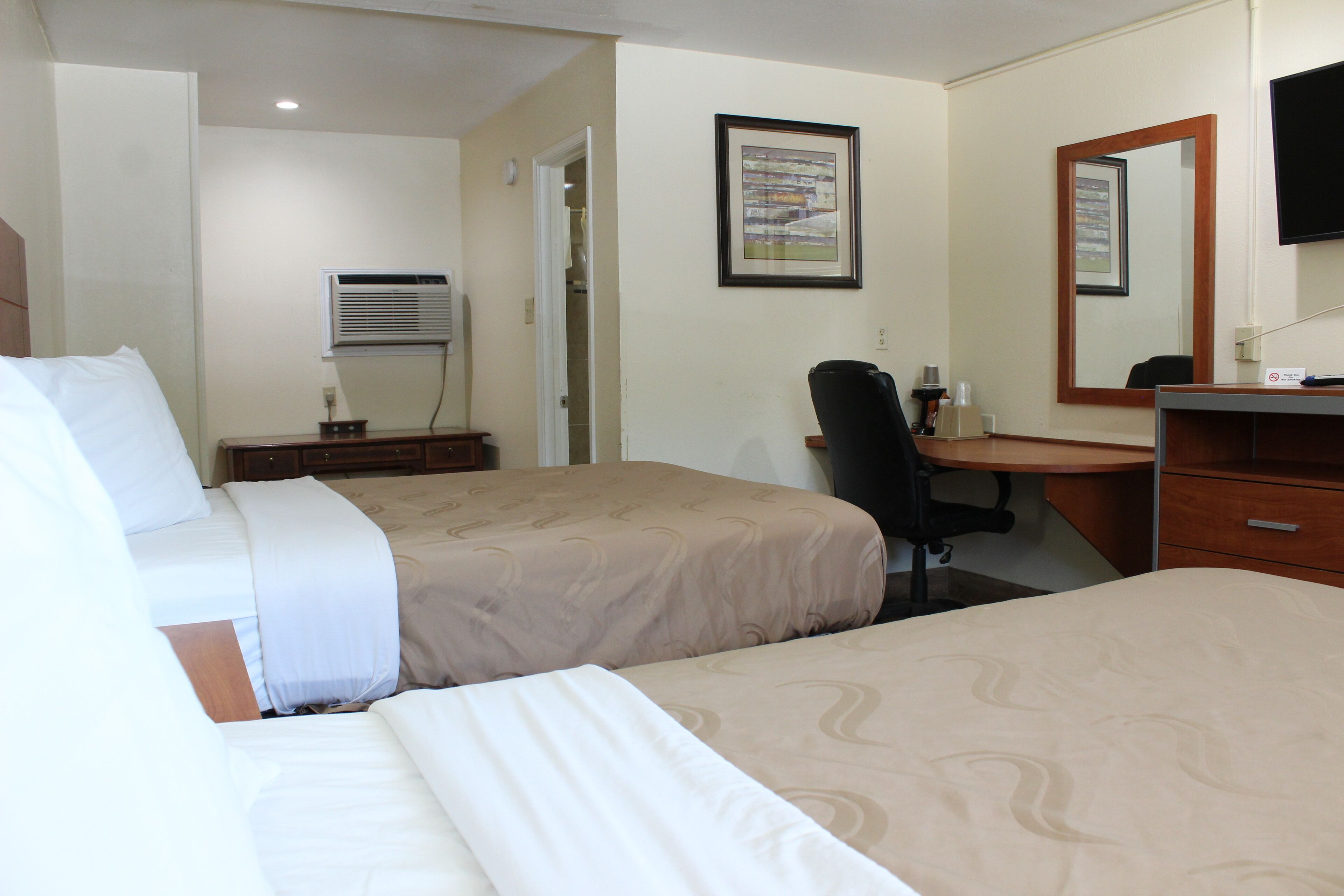 A hotel room with two beds, a desk, a chair, a mirror, and a wall-mounted TV.