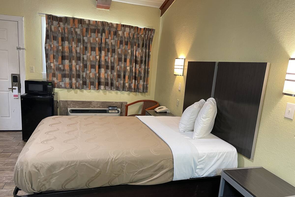 A hotel room with a bed, bedside table, a desk with a telephone, a microwave, and a heater.