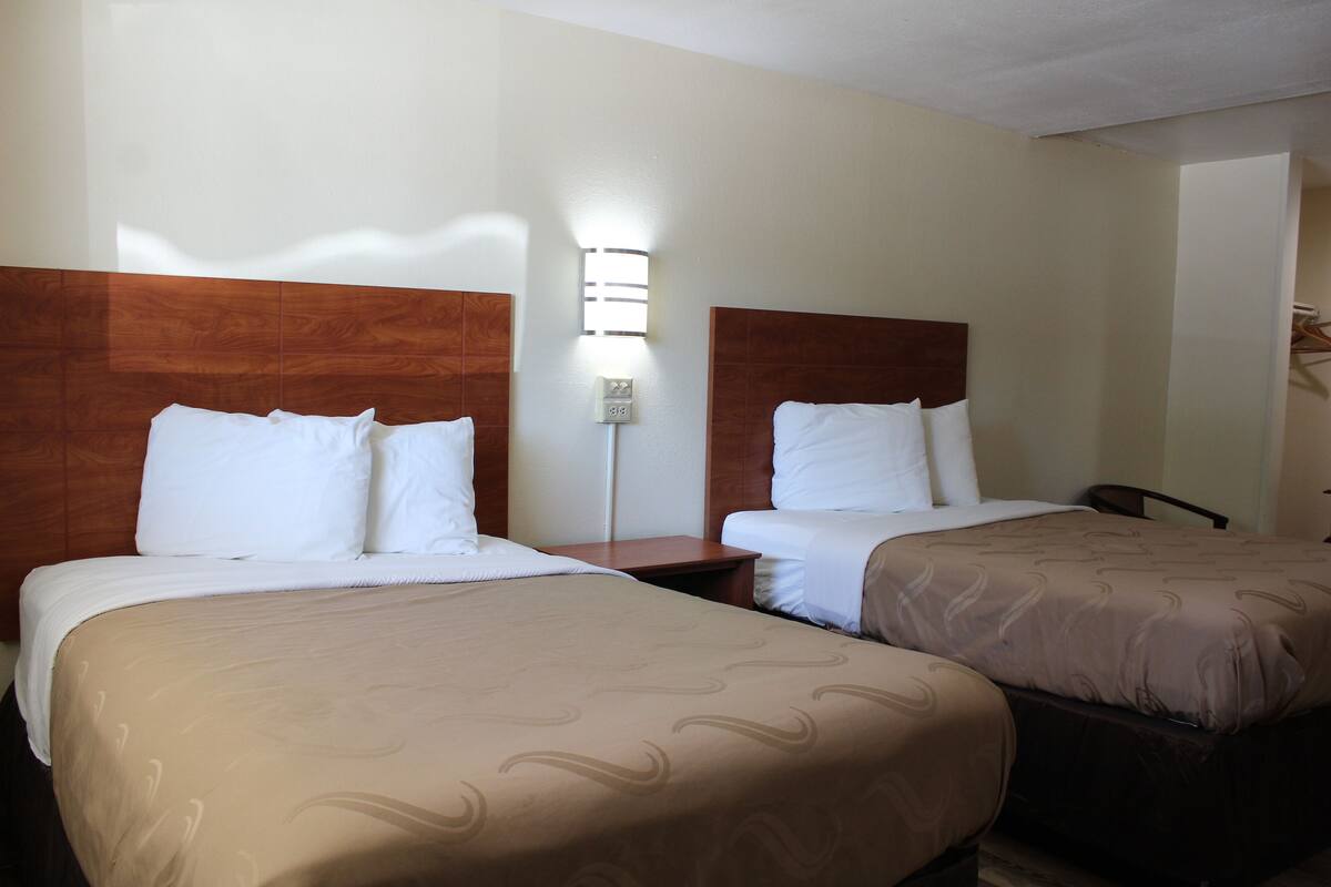 A hotel room with two beds, each with a wooden headboard and white bedding. There is a nightstand between the beds and a wall-mounted light fixture above it.