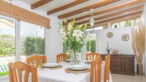 A dining area with a table set for six, wooden chairs, a vase with white flowers, and a view of the outdoors.
