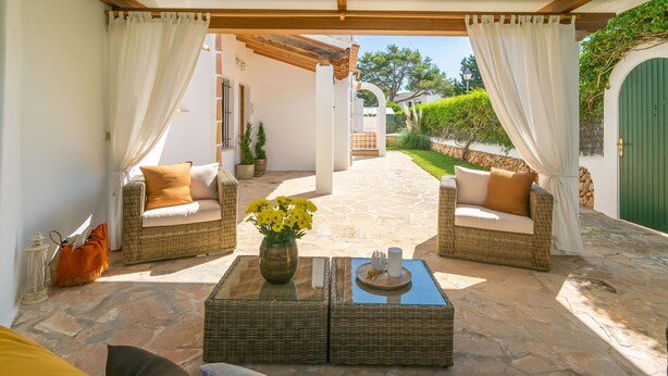 A covered outdoor seating area with wicker furniture and a central table with a vase of flowers.