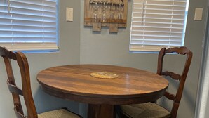 Una mesa de comedor redonda de madera con dos sillas tapizadas, junto a ventanas con persianas.