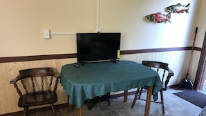 A room with a table covered by a teal tablecloth, two wooden chairs, a television on the table, and decorative fish on the wall.
