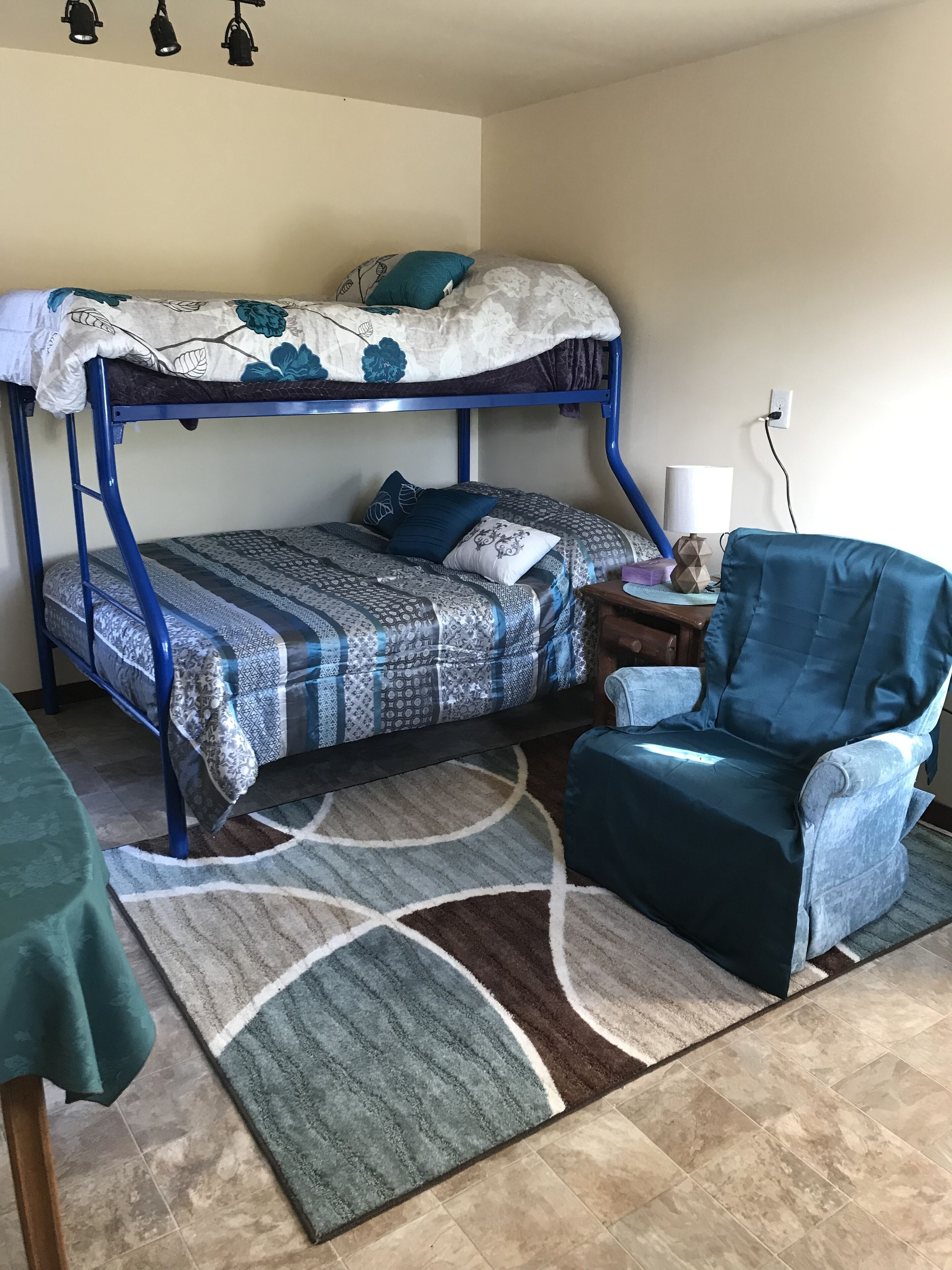 A bunk bed with patterned bedding, a blue chair, a lamp, and a rug in a room with tiled flooring.